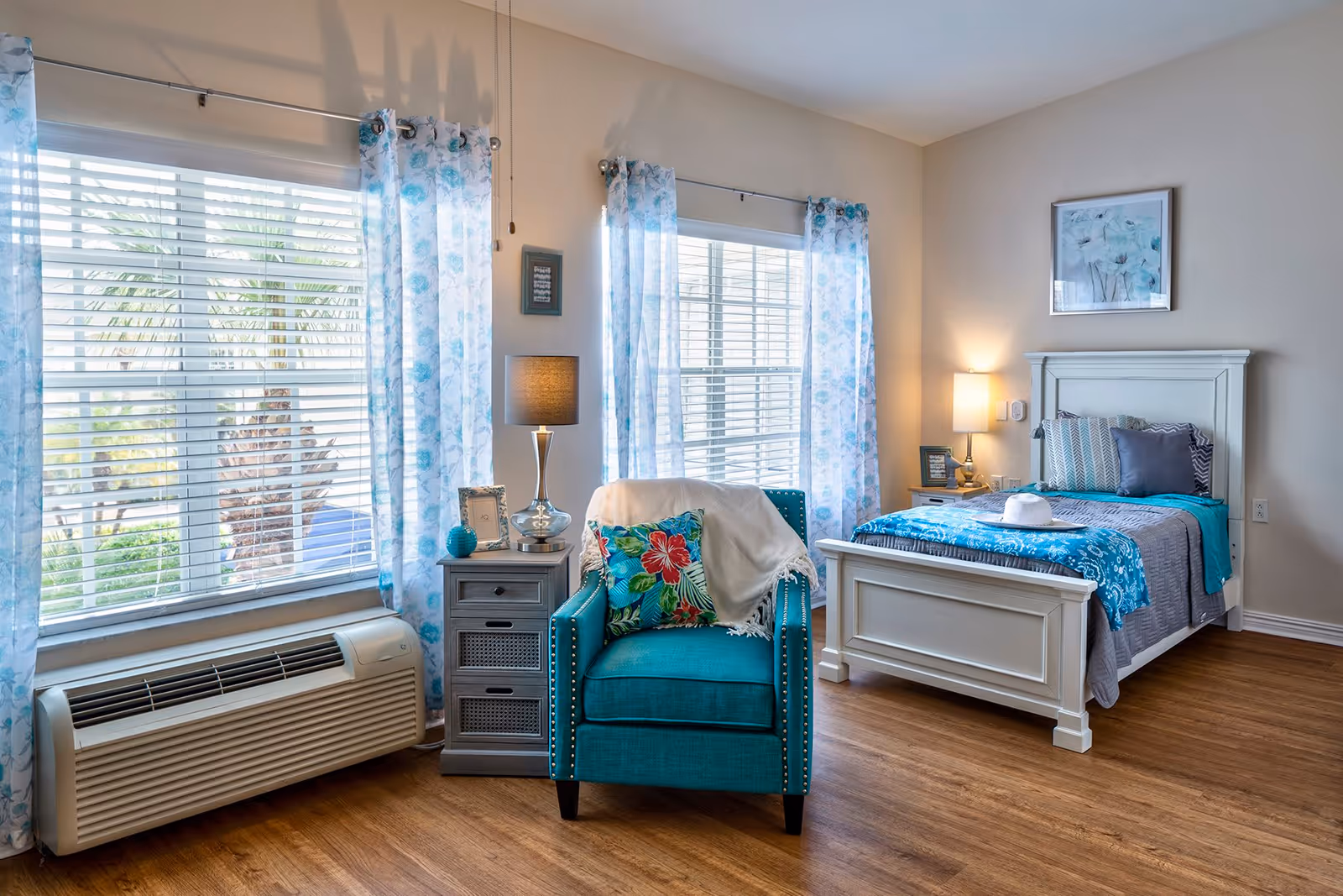 A bright and cozy bedroom featuring a white wooden bed with blue and gray bedding, a teal armchair with a floral pillow and a white throw, a gray nightstand with a lamp and decorative items, two large windows with white blinds and blue floral curtains, and wooden flooring.