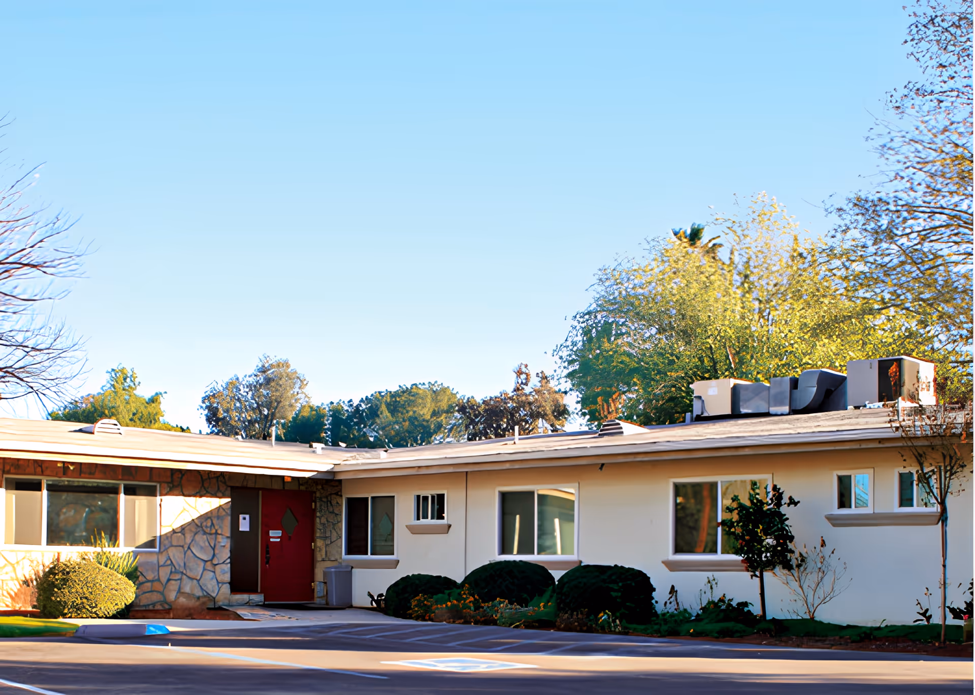 Exterior view of a single-story building with a red door, several windows, and bushes along the front. The building has a flat roof with HVAC units on top and is surrounded by trees under a clear blue sky.
