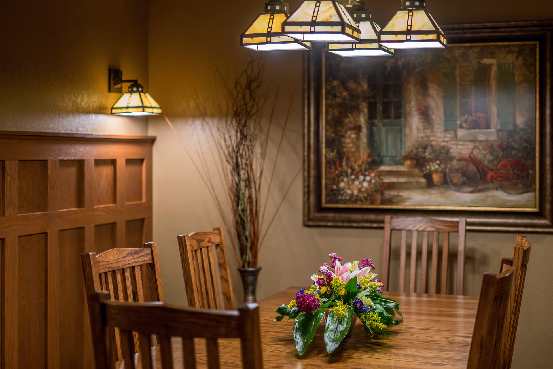 A warmly lit dining room with a wooden table and six wooden chairs. A floral centerpiece with colorful flowers is placed on the table. The walls are beige with wooden paneling on one side, and a large framed painting of a rustic outdoor scene with a bicycle hangs on the wall. A decorative vase with tall branches is in the corner, and three pendant lights with stained glass shades hang above the table.