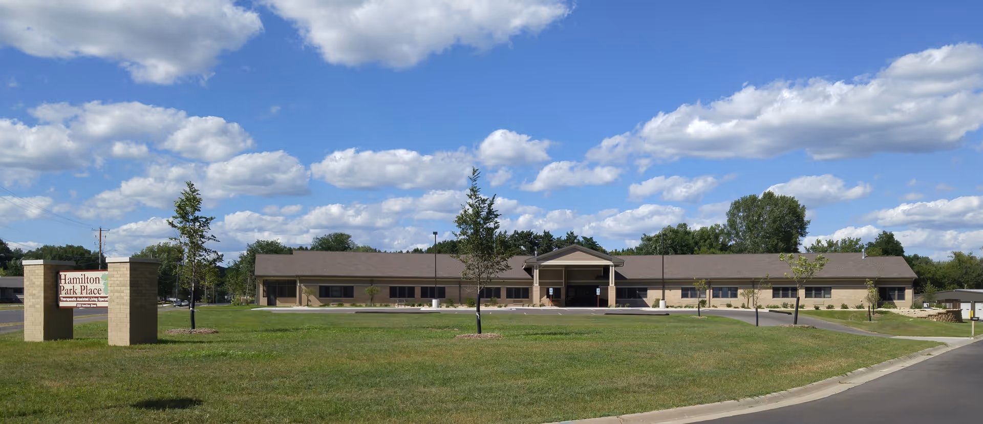 Front exterior of a single-story assisted living building with a lawn, entrance canopy, and a sign reading Hamilton Park Place under a blue sky.