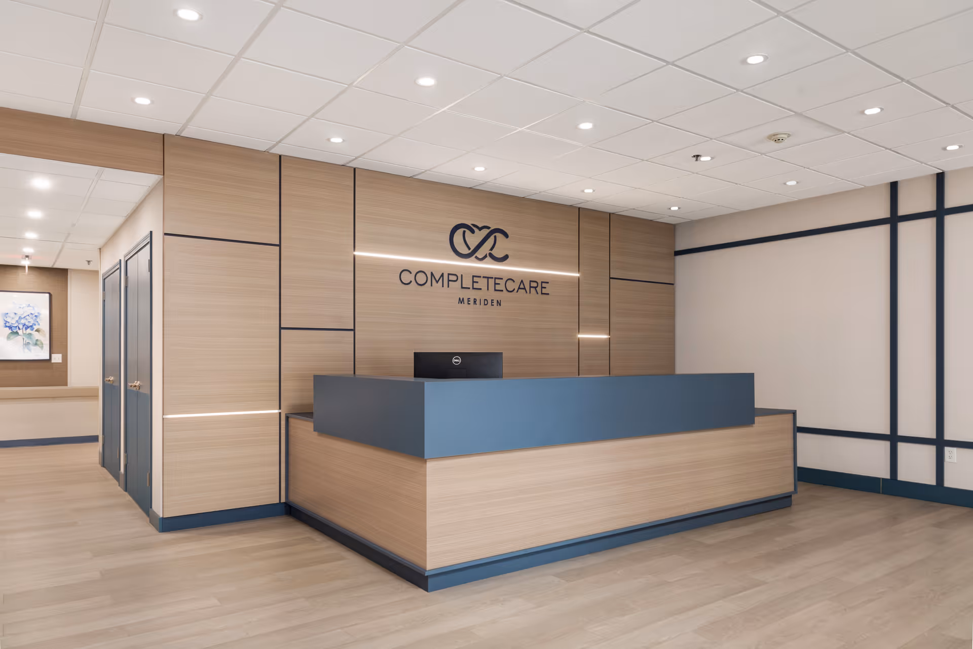 Reception area of Complete Care at Meriden featuring a modern wooden front desk with a blue countertop, light wood paneling on the walls, recessed ceiling lights, and the Complete Care Meriden logo displayed on the wall behind the desk.