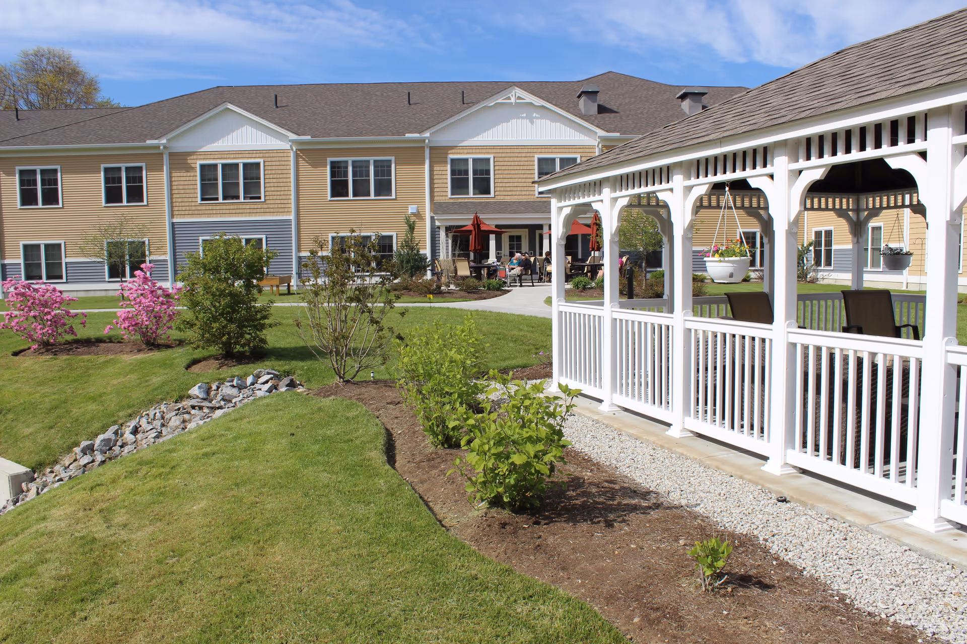 Outdoor view of a senior living facility with a well-maintained lawn, flowering bushes, a white gazebo with hanging flower pots, and a two-story building in the background under a blue sky.
