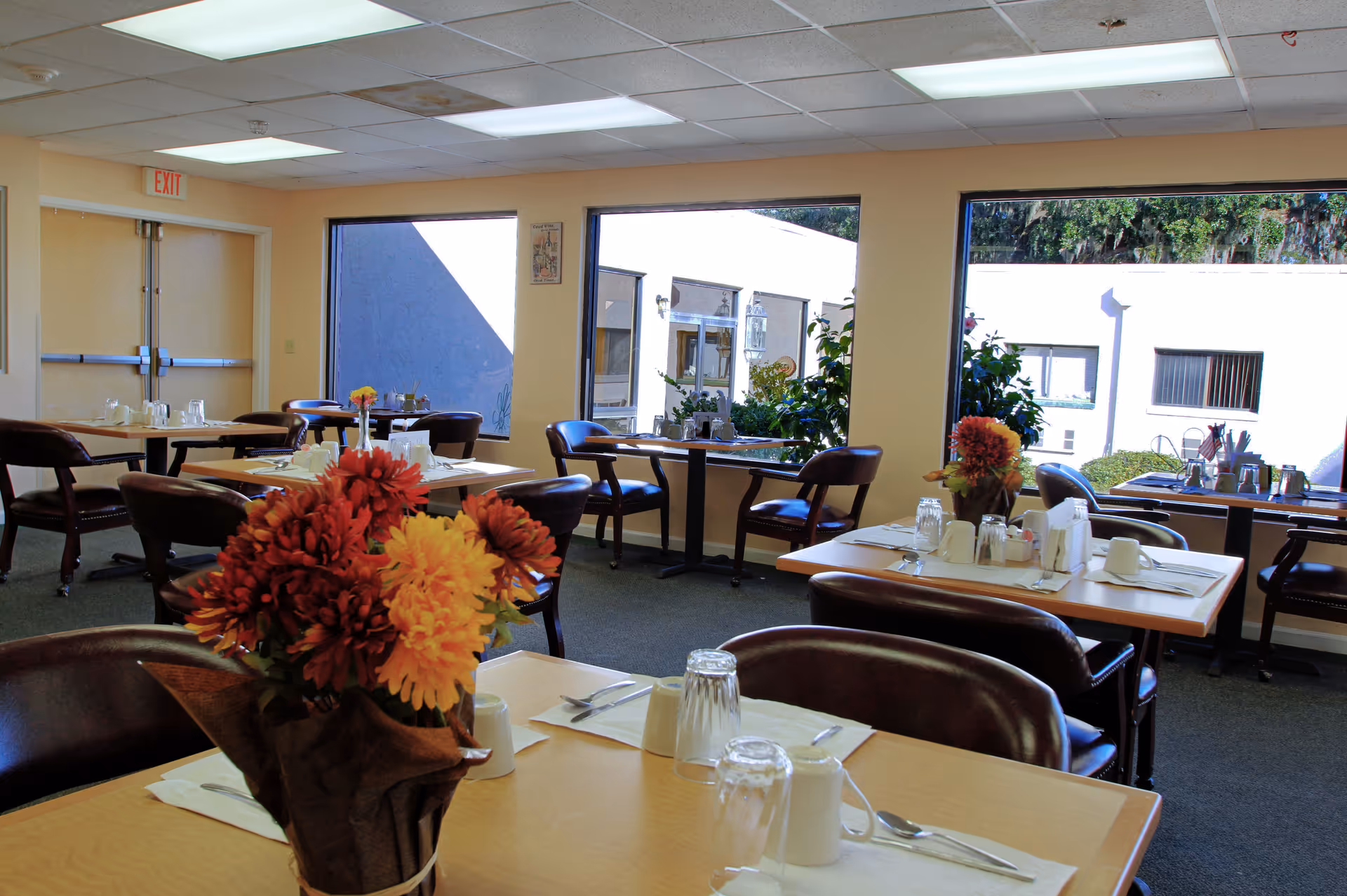 Dining room with set tables, chairs, and floral centerpieces beside large windows.