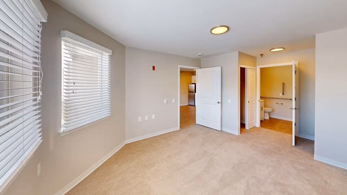Empty room with beige carpet and light gray walls featuring two windows with white blinds on the left wall. Two open doors on the far wall reveal a kitchen area with stainless steel appliances and a bathroom with a toilet and grab bars. Ceiling lights illuminate the space.