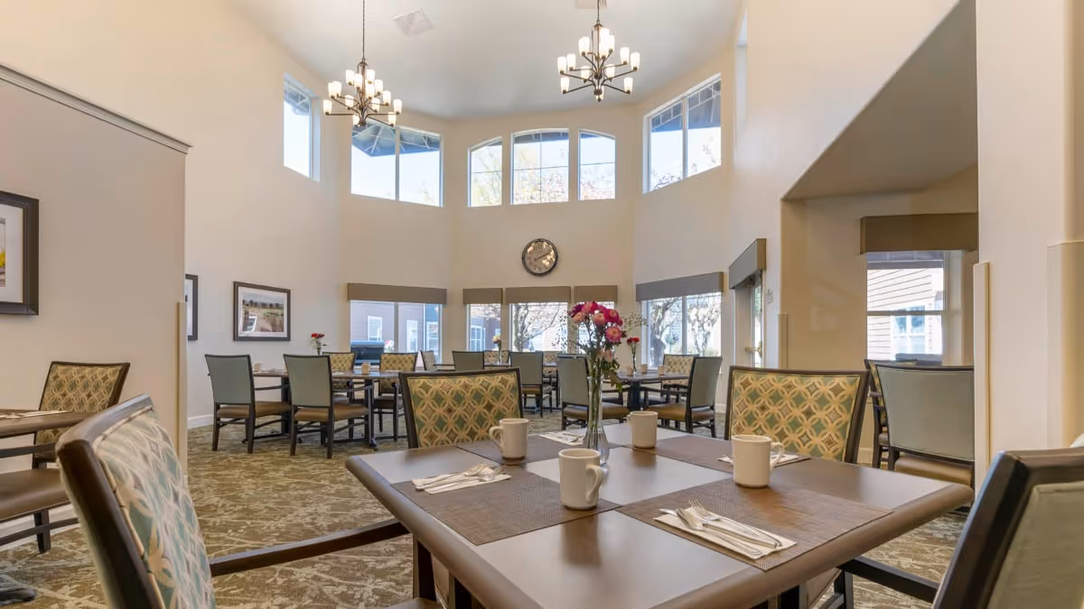 A bright dining room in a senior living facility with multiple tables and chairs arranged neatly. Each table has placemats, white mugs, and silverware. There are large windows letting in natural light, a clock on the wall, and two chandeliers hanging from the ceiling. The room has a warm and inviting atmosphere.