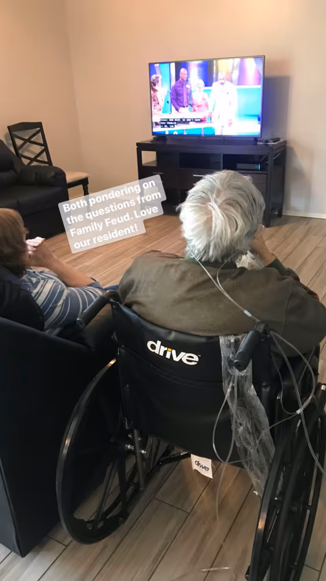 Two residents, one in a wheelchair, sit in a common room watching Family Feud on a TV.