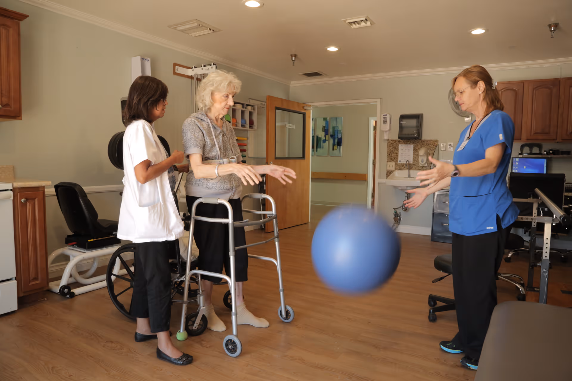 An elderly woman using a walker is engaged in a physical therapy exercise with two healthcare professionals in a therapy room. One professional is assisting the woman from behind, while the other is throwing a large blue exercise ball towards her. The room has wooden flooring, medical equipment, and cabinets.
