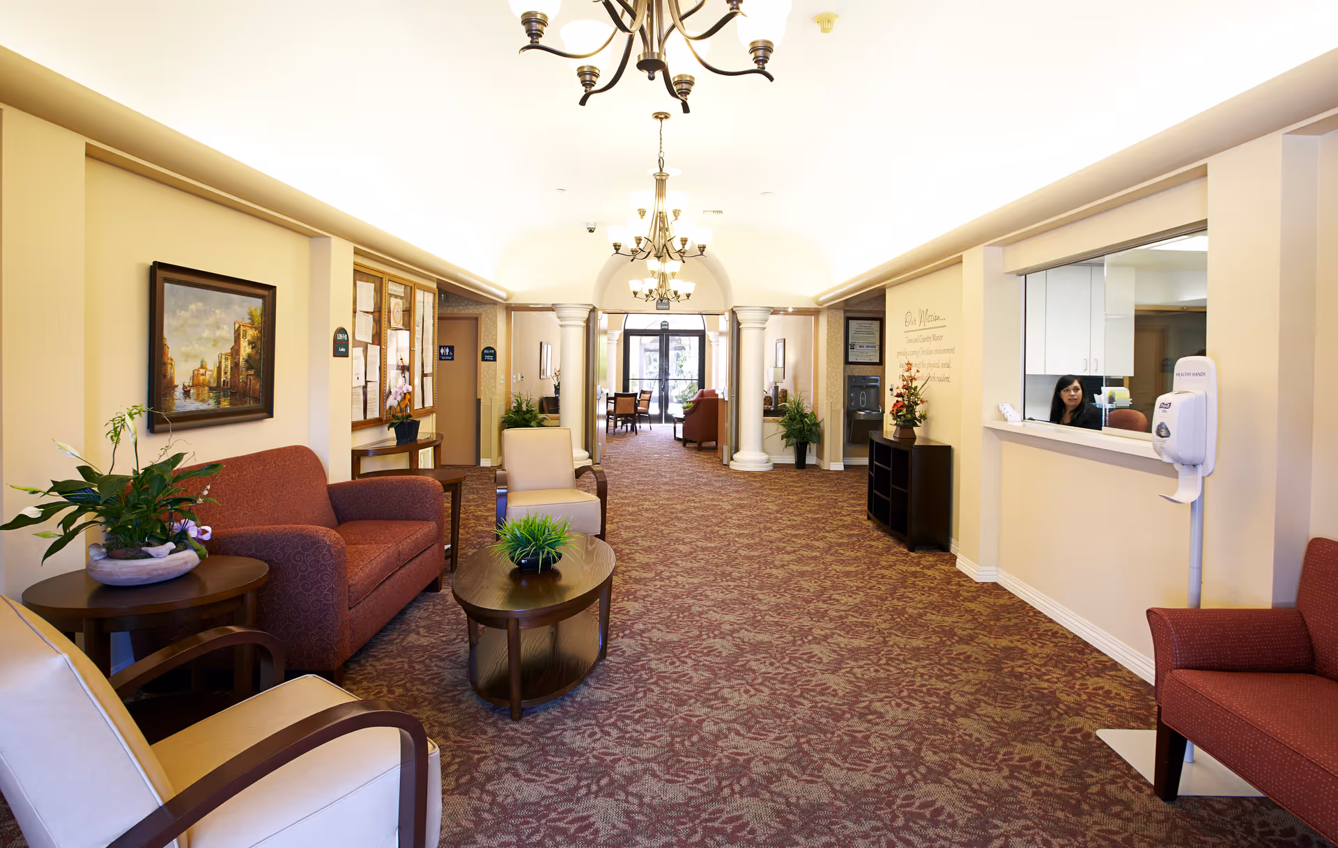 A spacious and well-lit senior living facility lobby with patterned carpet, beige walls, and multiple seating areas including armchairs and sofas. There are plants on tables, framed artwork on the walls, chandeliers hanging from the ceiling, and a reception window with a staff member visible behind it. The hallway leads to other rooms and an exit door with glass panels.