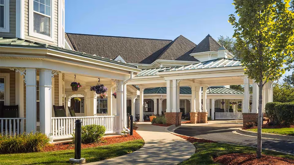 Exterior view of a senior living facility with a covered entrance, white columns, hanging flower baskets, and a curved walkway surrounded by landscaped greenery and trees under a clear blue sky.