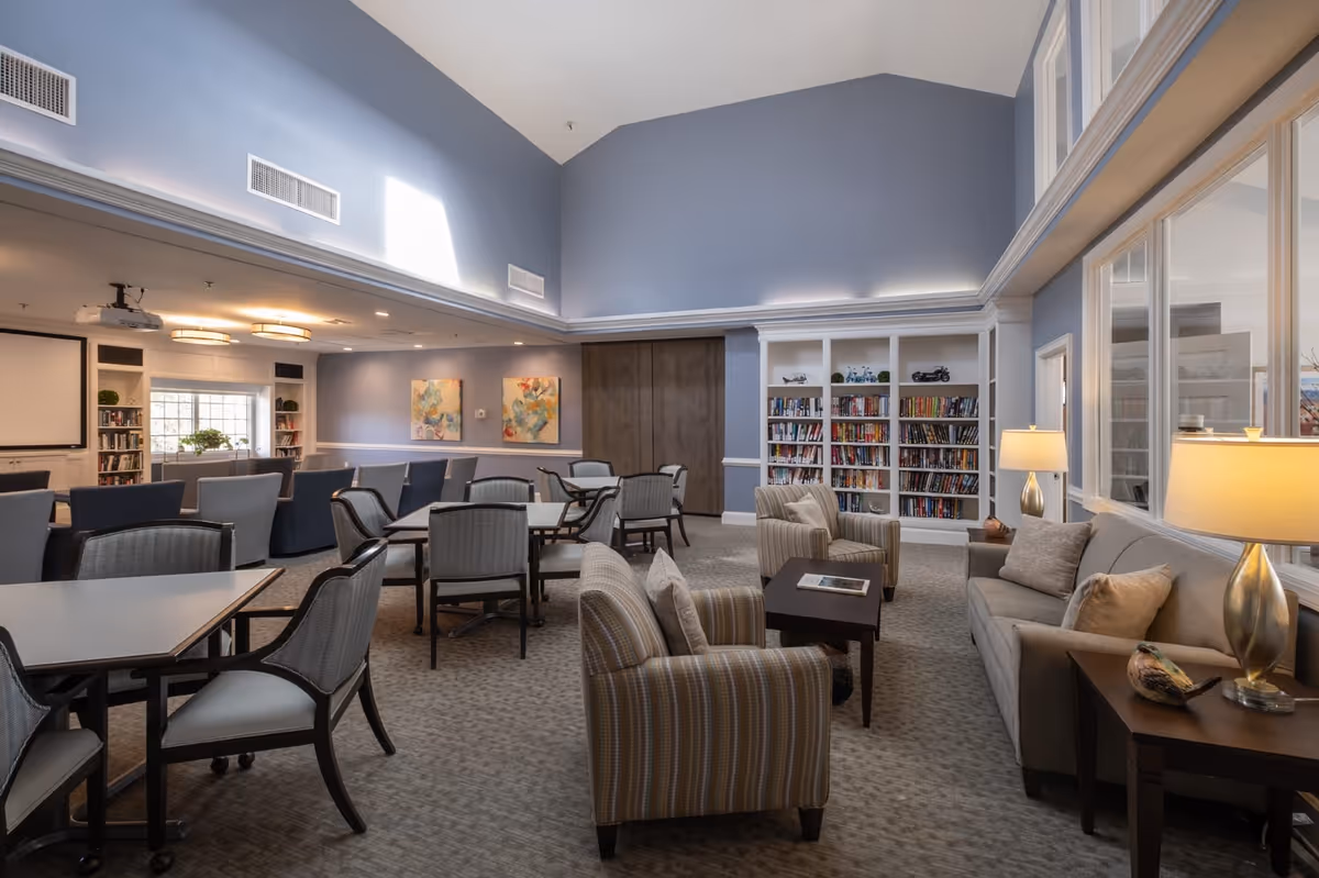 A spacious, well-lit common area with a high vaulted ceiling painted light blue. The room features several tables and chairs arranged for group seating, a cozy sitting area with a sofa, armchairs, and side tables with lamps. There are bookshelves filled with books along the back wall, two colorful abstract paintings, and a projector screen at the far end of the room.