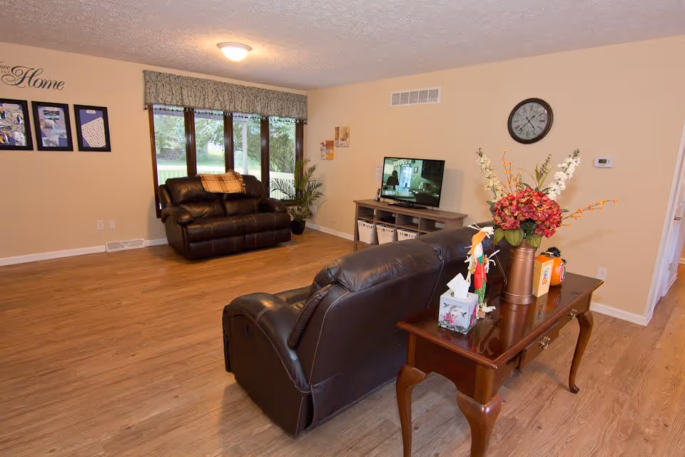 A cozy living room with wooden flooring, two dark brown leather sofas, a wooden side table with a flower vase and decorative items, a TV on a stand, a wall clock, and large windows with a valance letting in natural light.