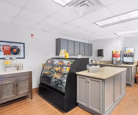 Interior view of a small food service area with a glass display case containing pastries and snacks, a countertop island with a basket, beverage dispensers in the background, gray cabinets, and a wooden sideboard with a framed picture on the wall.
