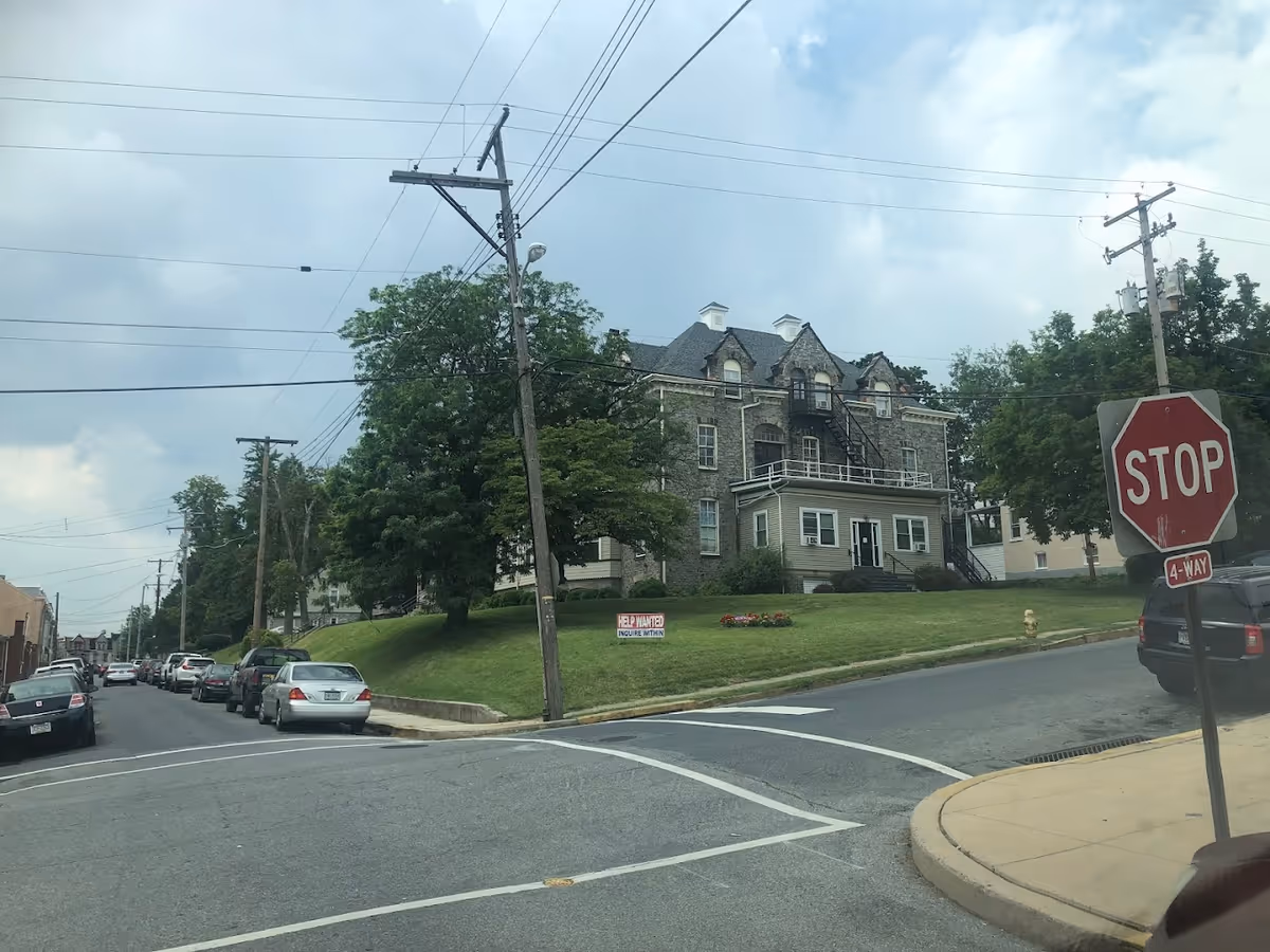 Stone multi-story house on a grassy corner lot with a stop sign and parked cars along the street.