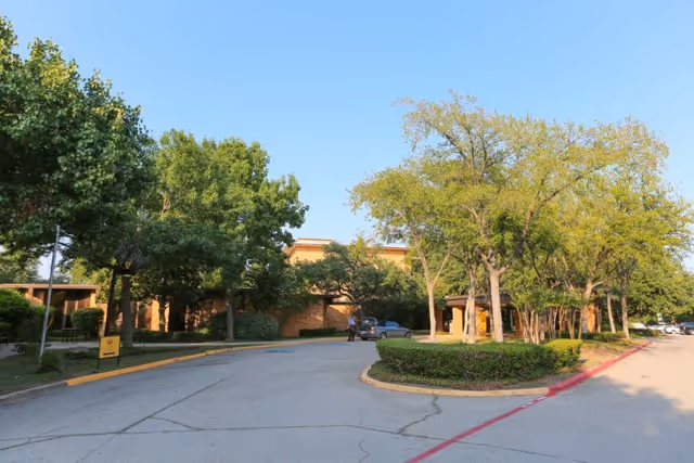 Exterior view of a senior living facility with a driveway, surrounded by green trees and bushes under a clear blue sky. A person is walking near a parked car close to the building entrance.