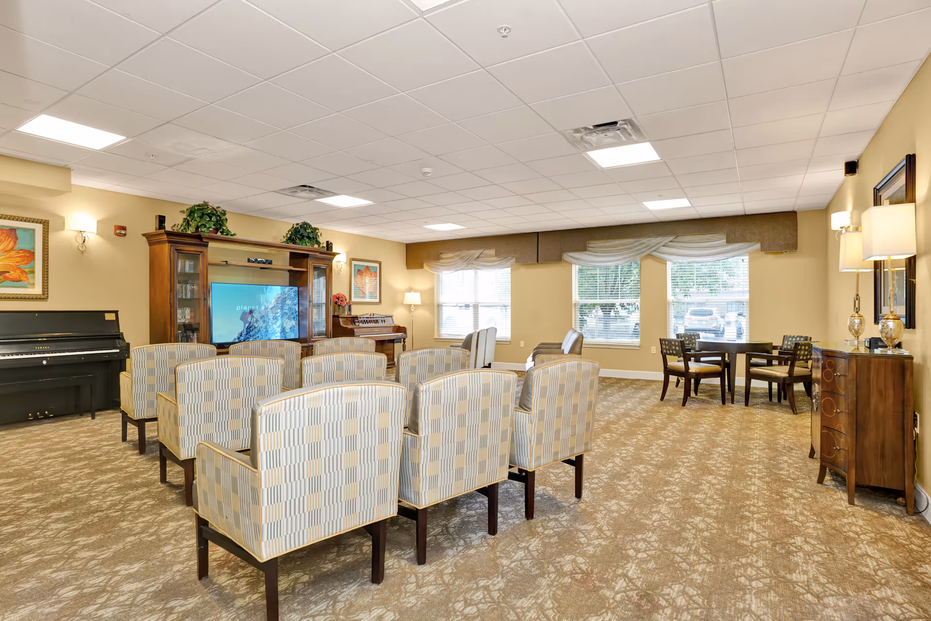 A spacious common area with multiple upholstered chairs arranged in rows facing a large wooden entertainment center with a TV. The room has beige walls, carpeted floor, and several windows letting in natural light. There is a piano on the left side and a small table with chairs near the windows on the right. Decorative plants and framed artwork adorn the walls.