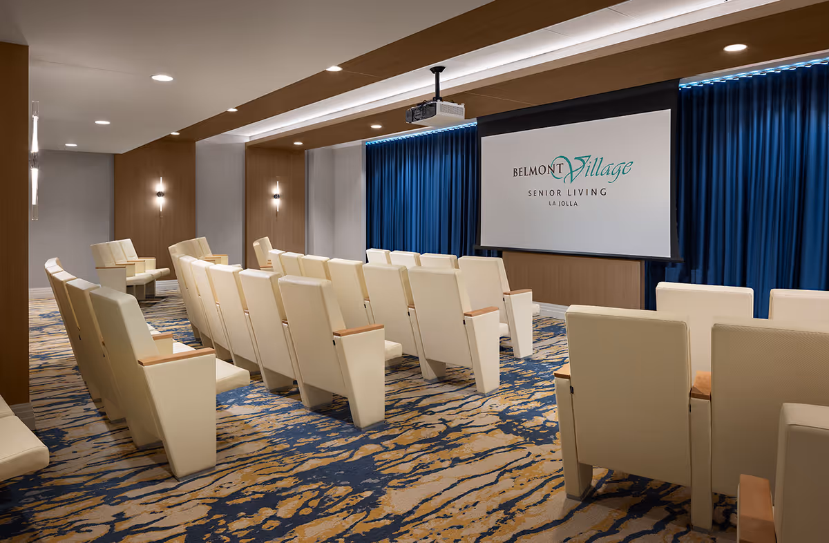 A modern presentation room with rows of beige cushioned chairs facing a large screen displaying the Belmont Village Senior Living La Jolla logo. The room features wood paneling, blue curtains behind the screen, recessed lighting, and a patterned carpet in shades of blue and beige.