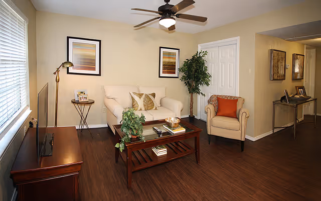 A cozy living room with a white loveseat and a beige armchair, both adorned with decorative pillows. A wooden coffee table with books, a plant, and a decorative bowl sits in front of the loveseat. There is a wooden TV stand with a flat-screen TV on the left side near a window with blinds. Two framed abstract artworks hang on the beige wall behind the loveseat. A tall floor lamp and a potted plant are also visible. The room has wooden flooring and a ceiling fan with lights. A hallway with a console table and framed pictures is visible to the right.