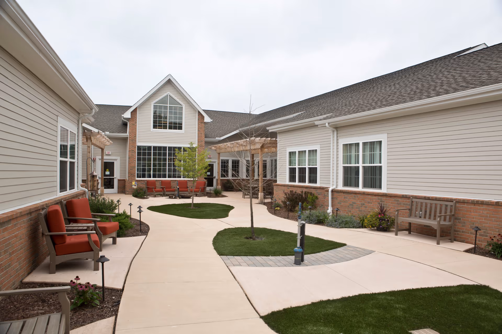 Outdoor courtyard area of a senior living facility with beige siding and brick exterior walls. The courtyard features a paved walkway, small patches of grass with young trees, and several benches and chairs with red cushions arranged along the sides. The sky is overcast.
