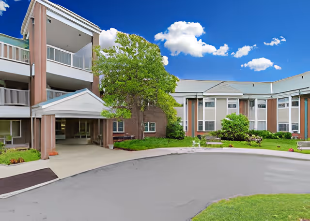 Front entrance and circular driveway of a multi-story assisted living building with balconies, a large tree, and landscaped lawn.