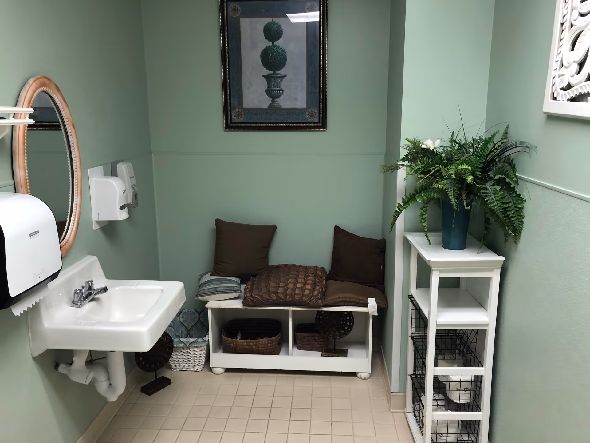 A small room with light green walls featuring a white wall-mounted sink with a paper towel dispenser and soap dispenser above it. There is a white bench with cushions and baskets underneath, a framed picture on the wall, and a white shelving unit with a green potted plant on top.