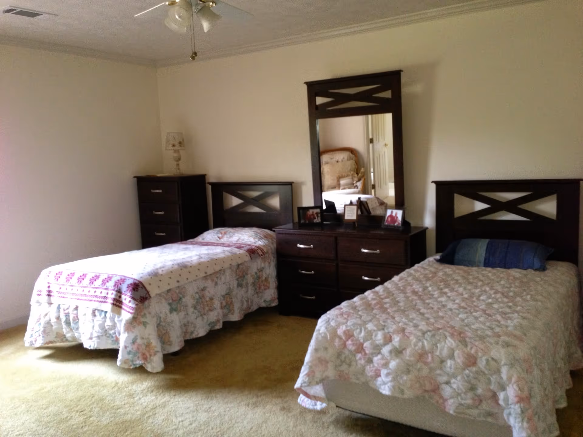A bedroom with two twin beds, a central dresser and mirror, a tall chest, and a lamp on light-colored walls and carpet.