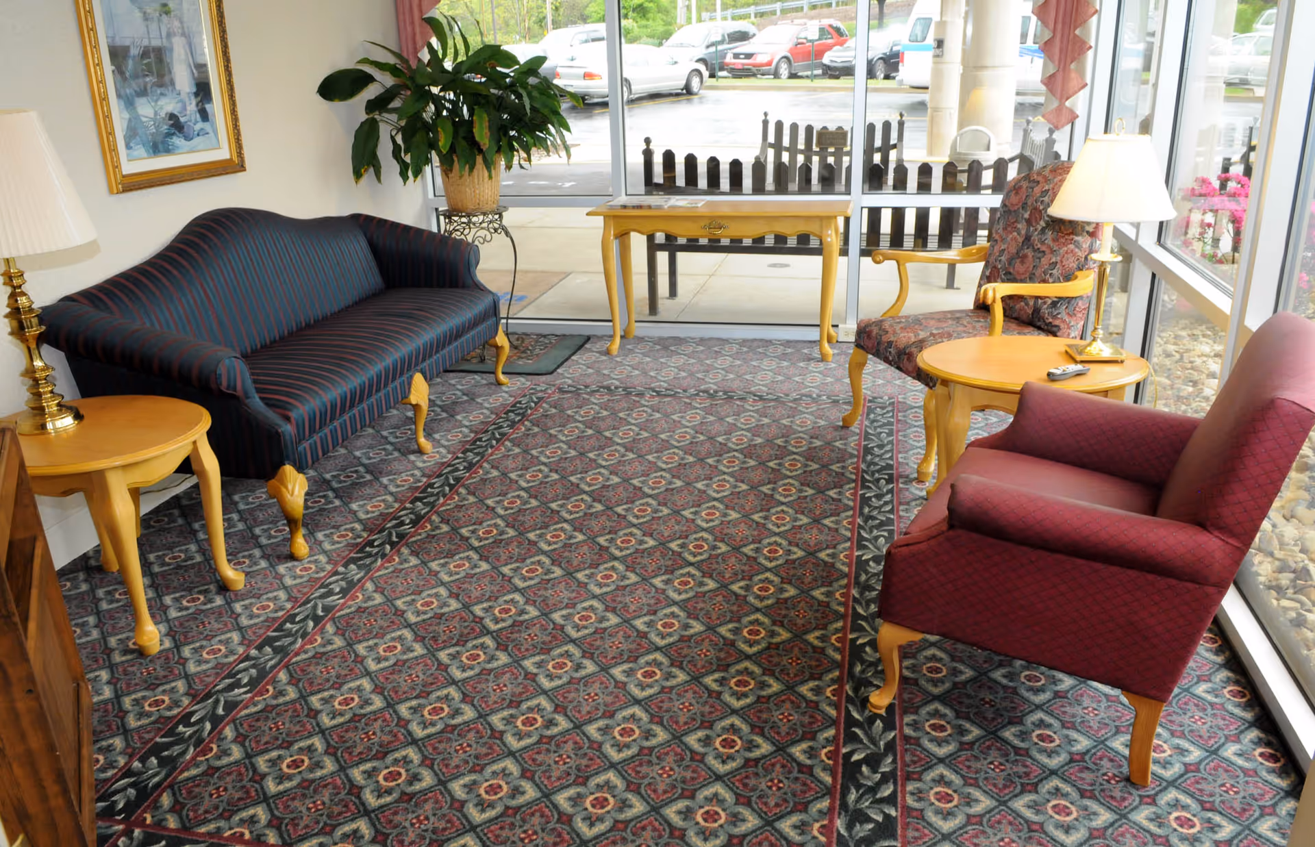 A cozy sitting area with a patterned carpet, a striped dark blue sofa, two armchairs with wooden legs and armrests, a small round wooden table with a lamp, a potted plant, and a wooden console table near large windows showing a parking lot outside.
