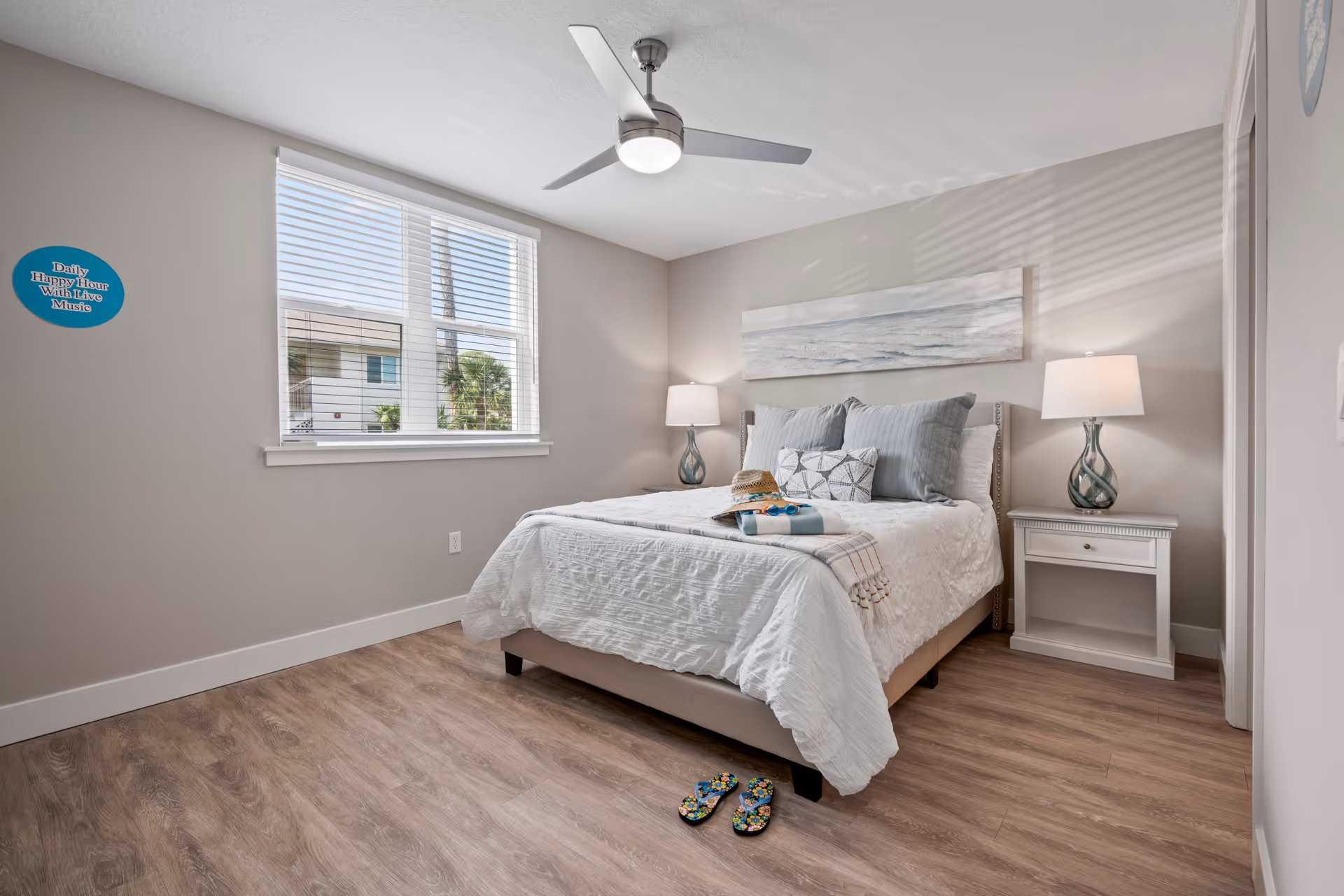 A bright bedroom with a large window letting in natural light. The room features a neatly made bed with white bedding and multiple pillows, two bedside tables each with a lamp, a ceiling fan with a light, and a piece of artwork above the bed depicting a serene ocean scene. On the bed are a hat, sunglasses, and a towel, and a pair of colorful flip-flops are placed on the wooden floor near the foot of the bed.