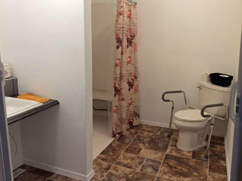 Bathroom with a toilet equipped with safety rails, a shower area with a floral patterned curtain, a sink with a countertop, and a tiled floor.
