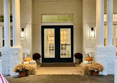Front entrance of a brick building with double glass-paned doors, columns, and autumn decorations including pumpkins, hay bales, and potted mums.