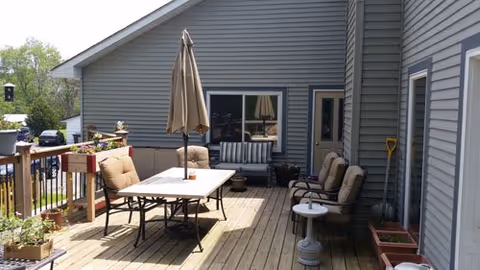Outdoor wooden deck with table, chairs, umbrella, and potted plants beside a gray building.