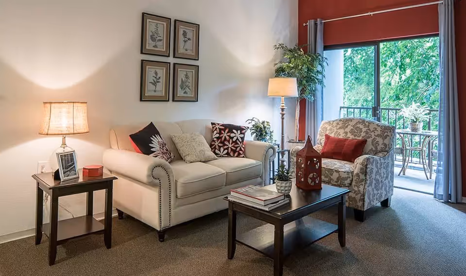 Cozy living room with a white sofa and patterned armchair around a coffee table, lamps, framed art, and a sliding glass door opening to a balcony.