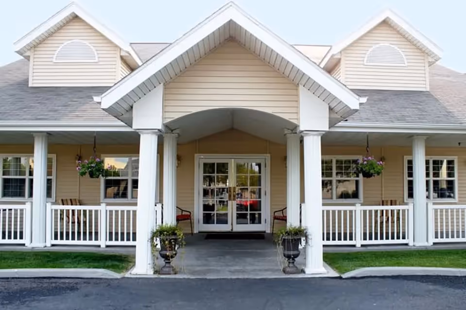 Front exterior view of a senior living facility with beige siding, white columns, a covered porch with hanging flower baskets, and glass double doors entrance.