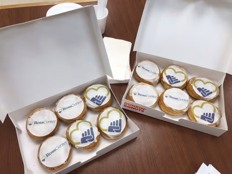 Two open boxes of donuts on a wooden table. Each donut has white icing with printed edible logos: some say 'Ross Center' and others show a blue hand inside a green heart. There are napkins and plastic utensils in a white cup behind the boxes.
