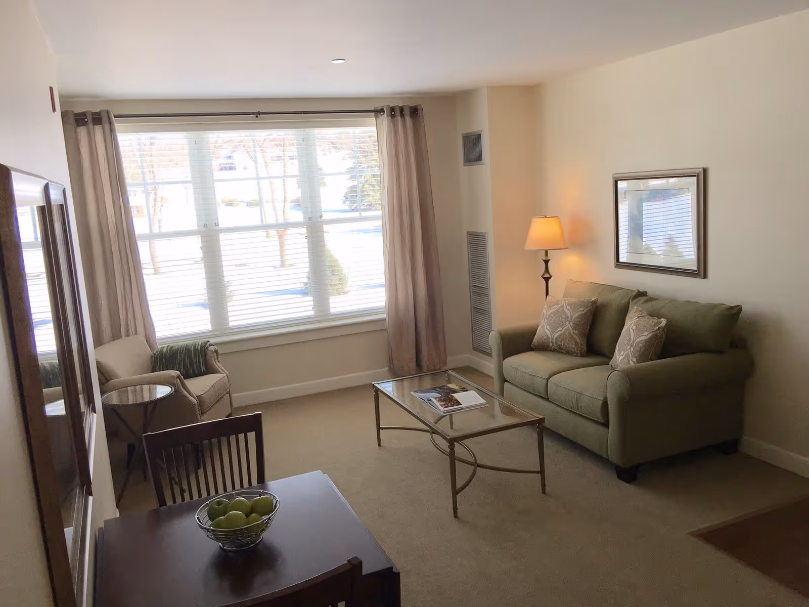 A cozy living room with a green loveseat adorned with two patterned pillows, a glass coffee table with a magazine on it, a floor lamp, and a framed picture on the wall. There is a large window with beige curtains letting in natural light, a beige armchair with a green throw blanket, and a small dining table with a bowl of green apples.