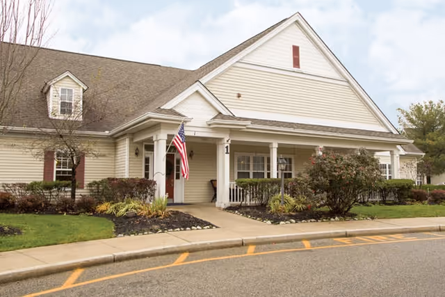 Single-story beige senior living building with a covered porch, American flag, and landscaped front entrance.