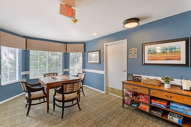 Small activity room with a square table and four chairs by bay windows, blue walls, and a console shelf stocked with board games and decor.
