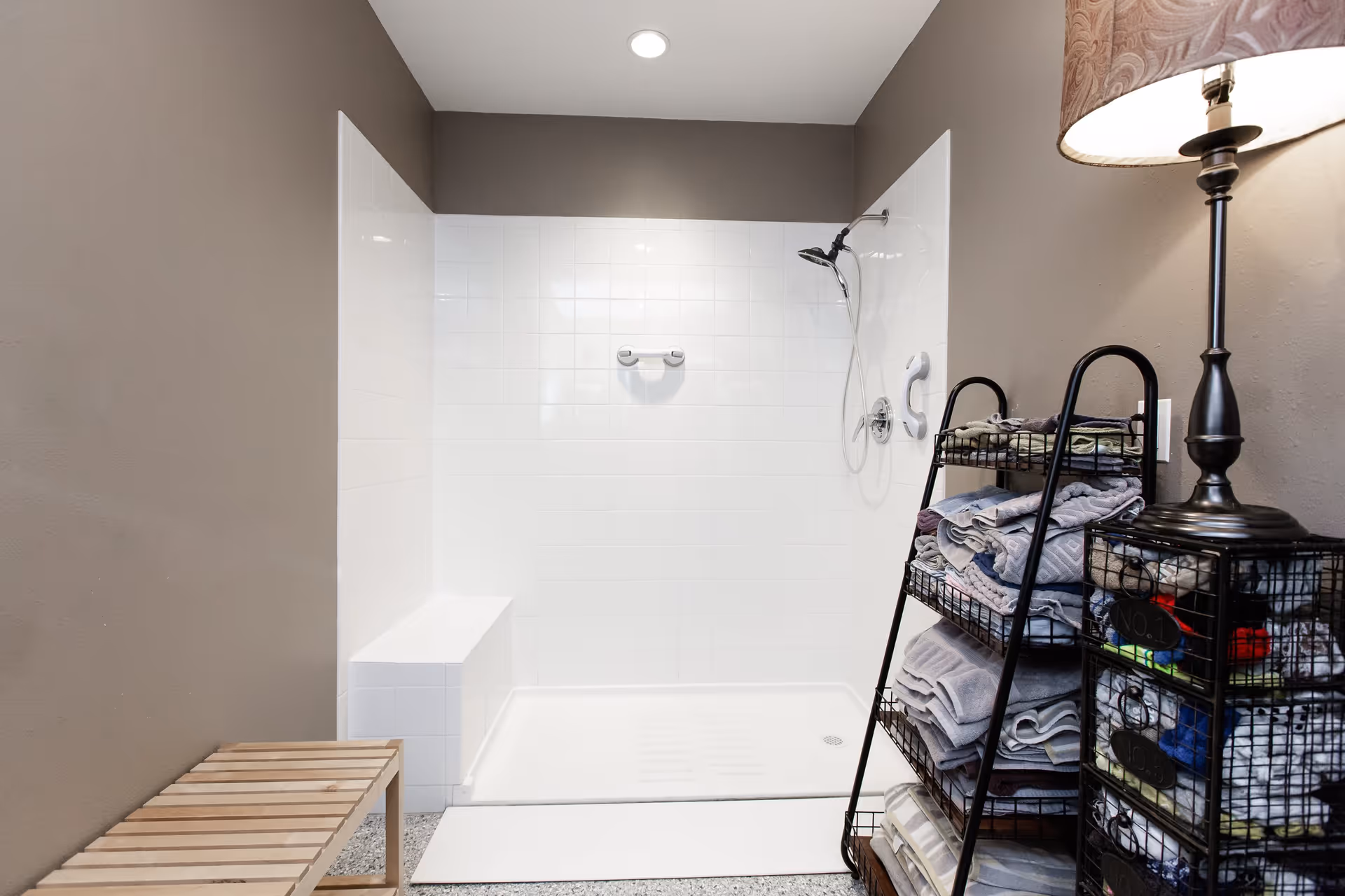 A white tiled walk-in shower with a built-in bench, handheld showerhead, and a shelving unit stacked with folded towels and a lamp nearby.