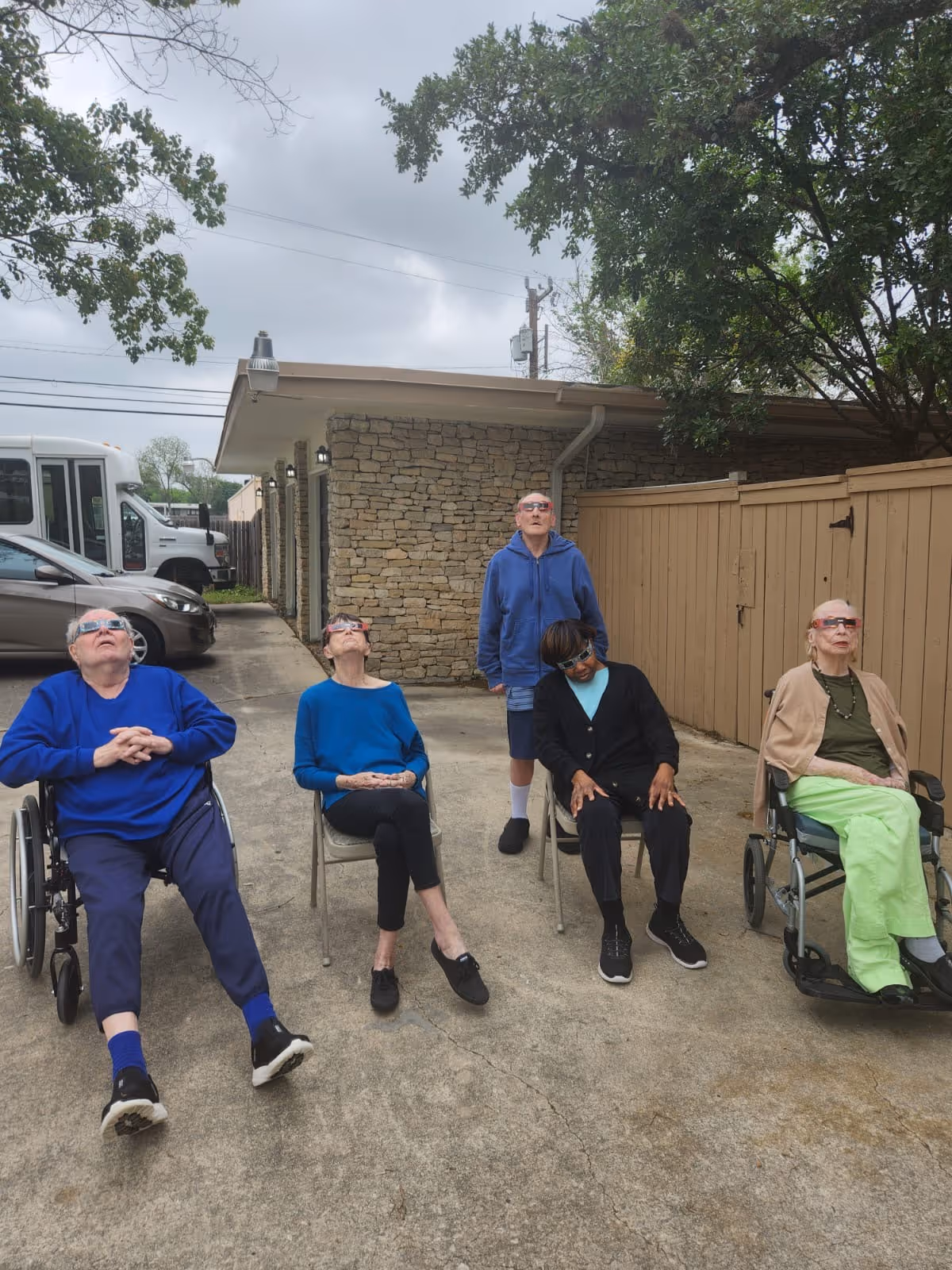 Five elderly individuals outdoors sitting and standing on a concrete surface near a stone building and wooden fence, all wearing protective glasses and looking up at the sky, possibly observing a solar eclipse or similar event.