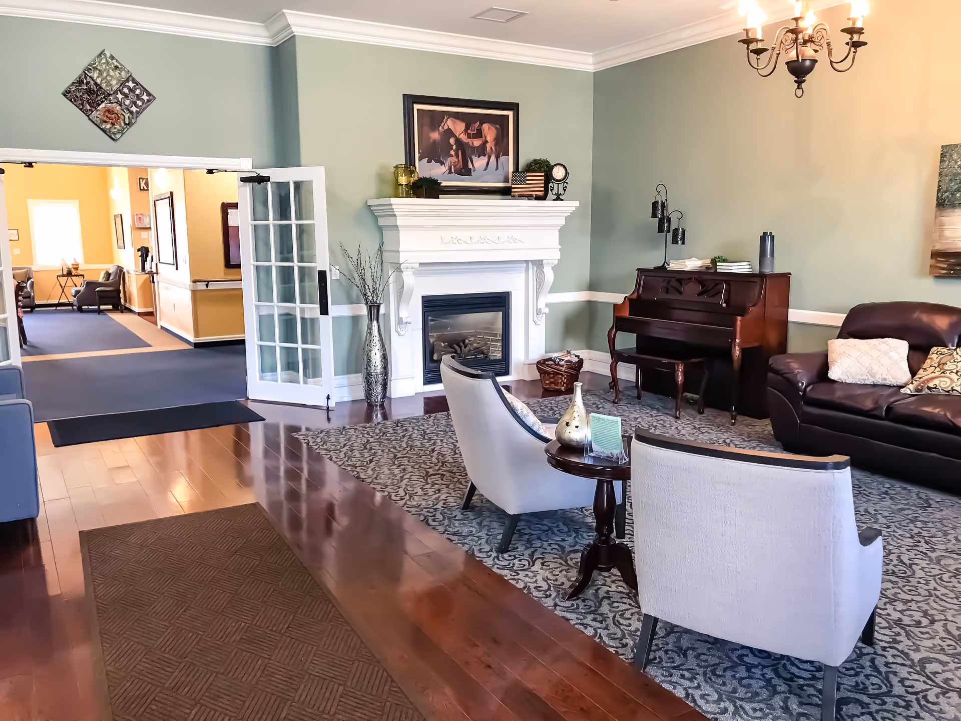 A cozy living room area in Tambree Meadows Assisted Living featuring a white fireplace with decorative items on the mantel, a dark wooden piano with a bench, a dark leather sofa with pillows, two light-colored armchairs around a small round table, a patterned area rug, hardwood floors, and a chandelier. French doors open to a hallway with additional seating visible.