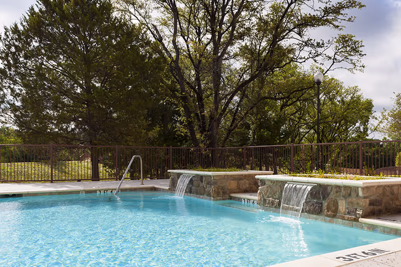 Outdoor swimming pool with two stone waterfall spouts, a metal handrail, and trees beyond a fence.