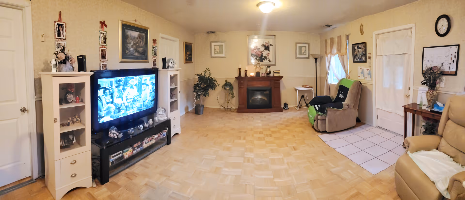 Spacious communal living room with a large TV and shelving on the left, an electric fireplace in the center, and recliner chairs on the right over parquet flooring.
