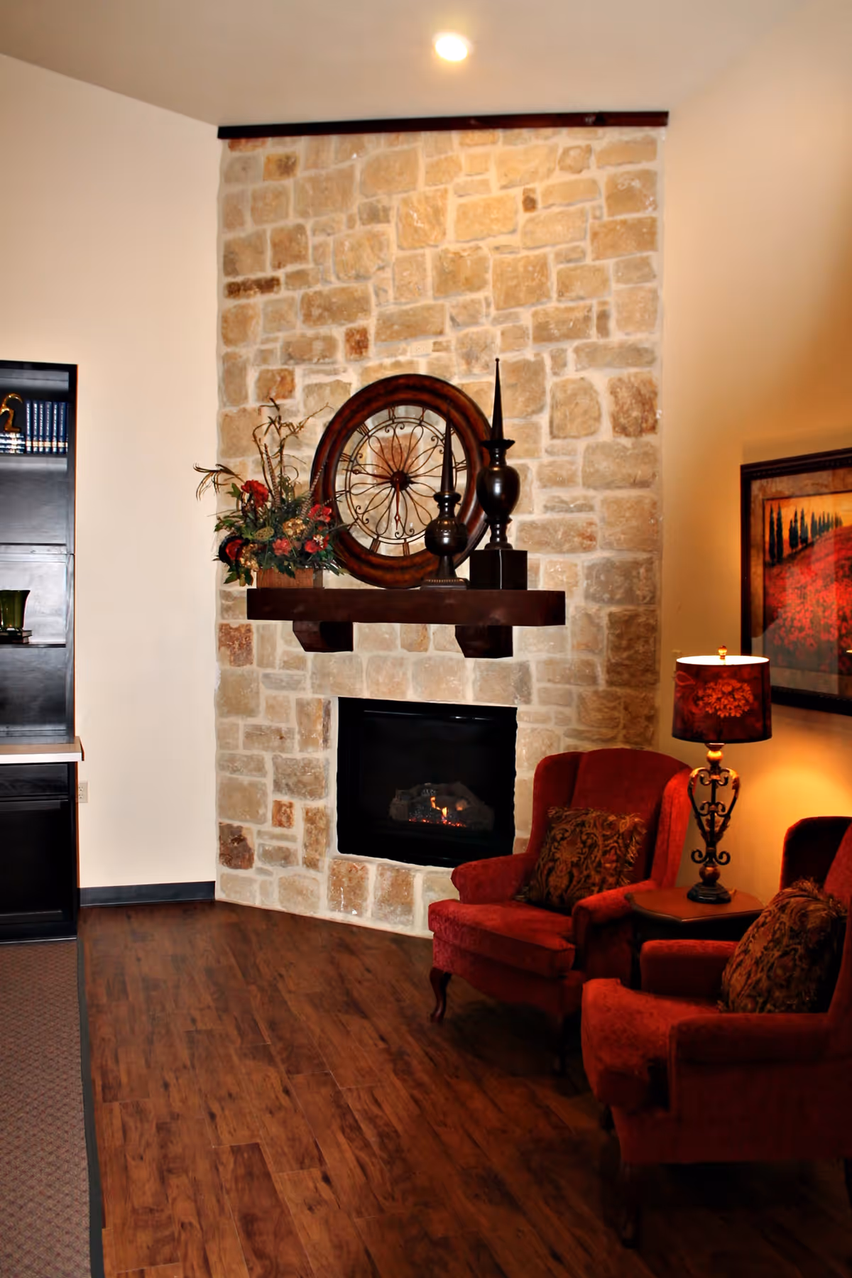 Cozy living room area with a stone fireplace, a wooden mantel decorated with a large round clock, vases, and a floral arrangement. Two red upholstered armchairs with patterned cushions are placed near a small side table with a decorative lamp. A framed painting hangs on the wall beside the chairs.