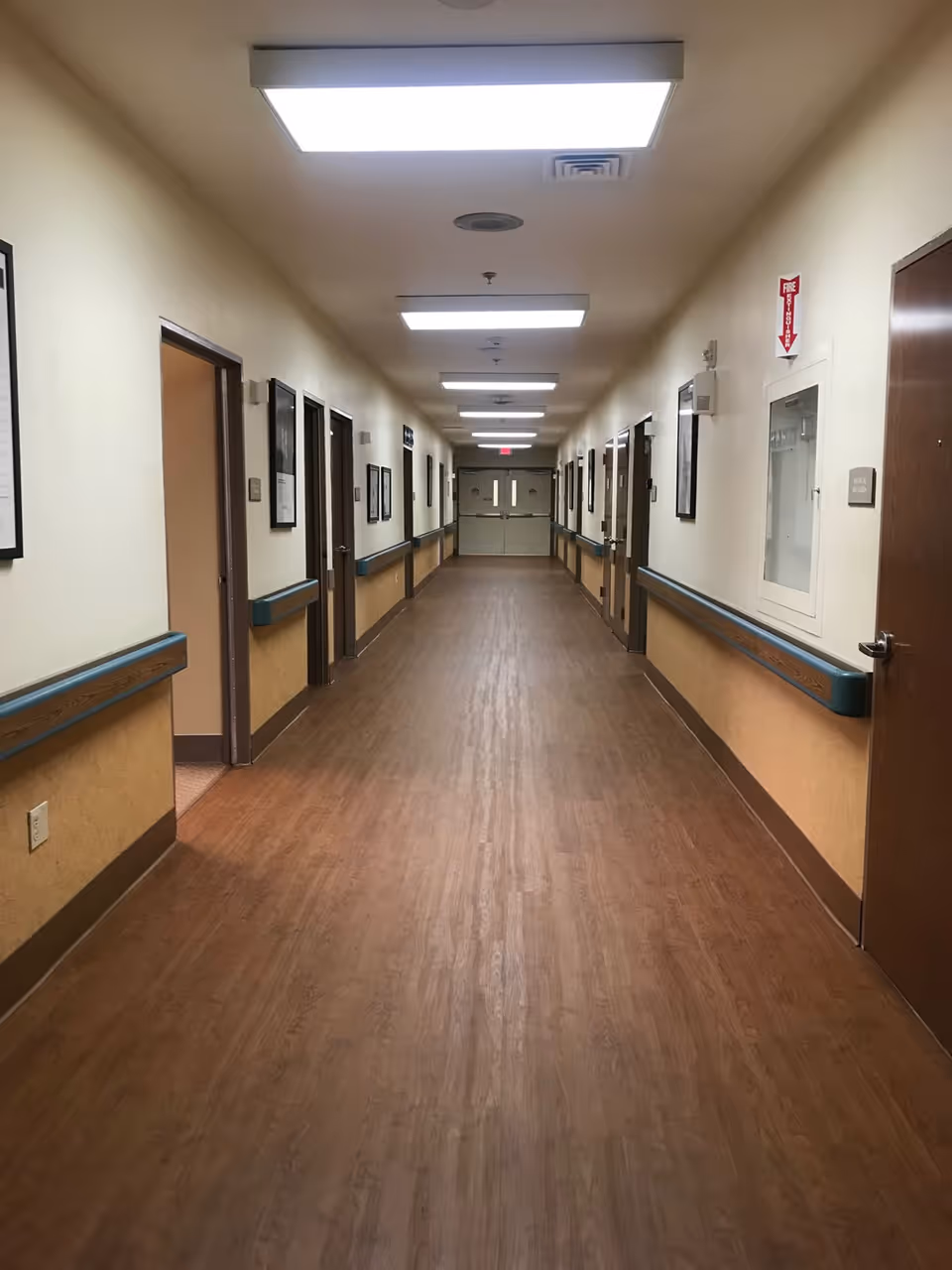 A long, well-lit hallway in a senior care facility with wood-patterned flooring, beige walls, handrails on both sides, multiple doors along the corridor, framed pictures on the walls, and double exit doors at the far end.