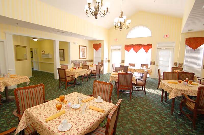 A bright dining room with multiple tables covered in patterned tablecloths and set with glasses, cups, and napkins. The room has green carpet, striped upholstered chairs, yellow walls with white trim, and large windows with red valances letting in natural light. Two chandeliers hang from the ceiling.