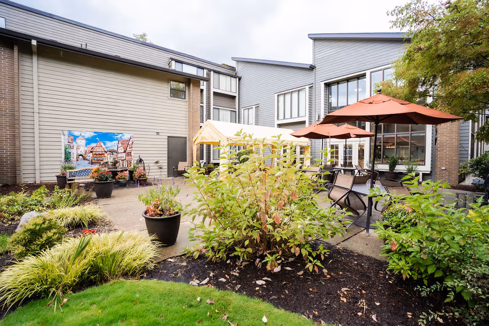 Outdoor patio area at Creekside Village Retirement Residence featuring several tables with orange umbrellas, surrounded by green plants and shrubs. The building exterior is visible with large windows and beige siding. There is a mural on the wall depicting a scenic village.
