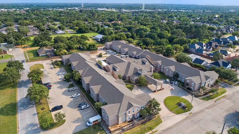 Aerial view of Morada Pantego senior living facility showing multiple connected buildings with gray roofs, surrounded by parking lots, trees, and nearby residential houses under a clear sky.