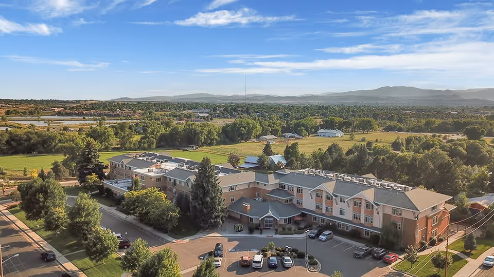 Aerial view of Hillcrest Of Loveland, a large multi-story senior living facility surrounded by trees, parking spaces with cars, and open green fields extending into the distance with mountains on the horizon under a partly cloudy sky.