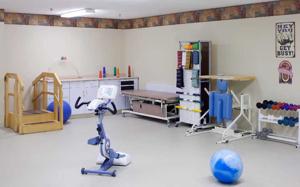 A physical therapy or rehabilitation room with exercise equipment including a stationary bike, wooden stairs with handrails, therapy table, exercise balls, resistance bands, dumbbells, and a motivational poster on the wall that says 'HEY YOU GET BUSY!'