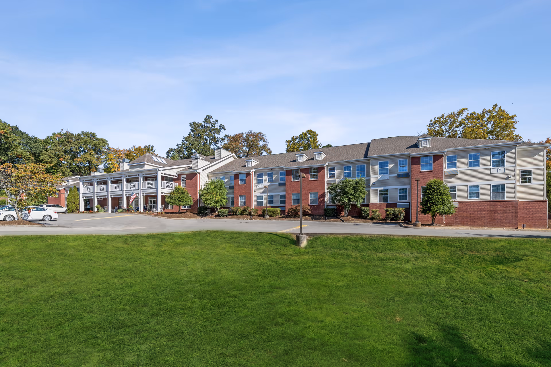 Exterior view of a multi-story senior living facility building with a combination of brick and siding. The building has multiple windows and a covered entrance with white columns. There are several trees and shrubs around the building, a paved driveway, parked cars, and a large green lawn in the foreground under a clear blue sky.