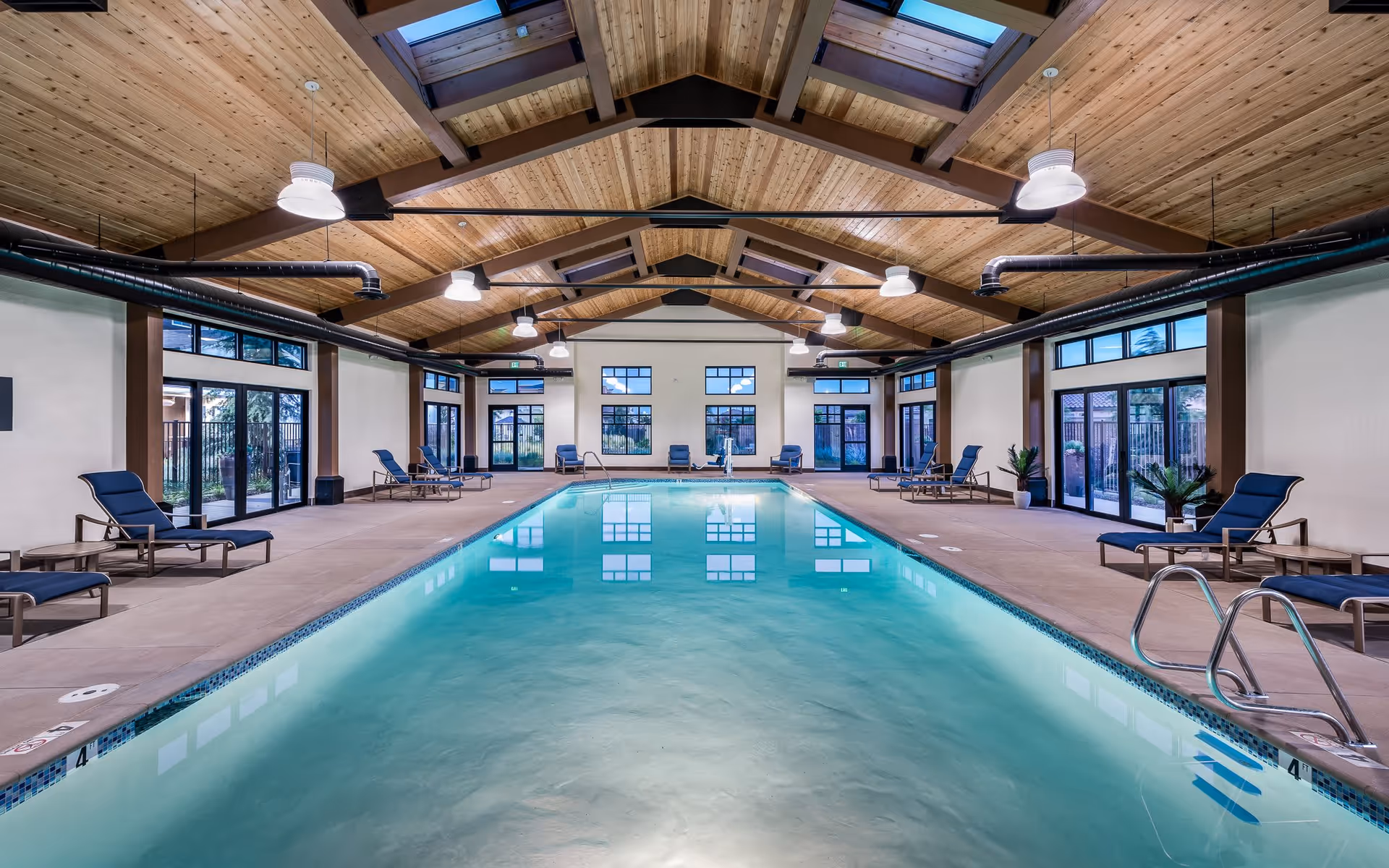 Indoor swimming pool with clear water, surrounded by lounge chairs and tables. The room has a high wooden ceiling with exposed beams and skylights, large windows and glass doors allowing natural light to enter.